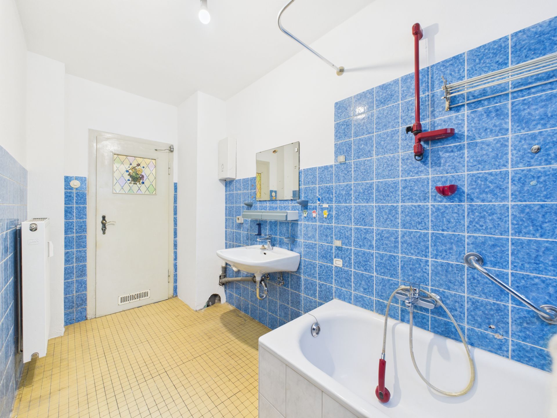 Badezimmer mit blauen Fliesen, Badewanne, Waschbecken und Spiegel, gelber Fliesenboden.