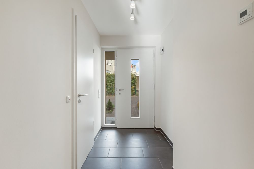 Heller Flur mit weißer Tür, Seitenfenster und dunklem Fliesenboden.