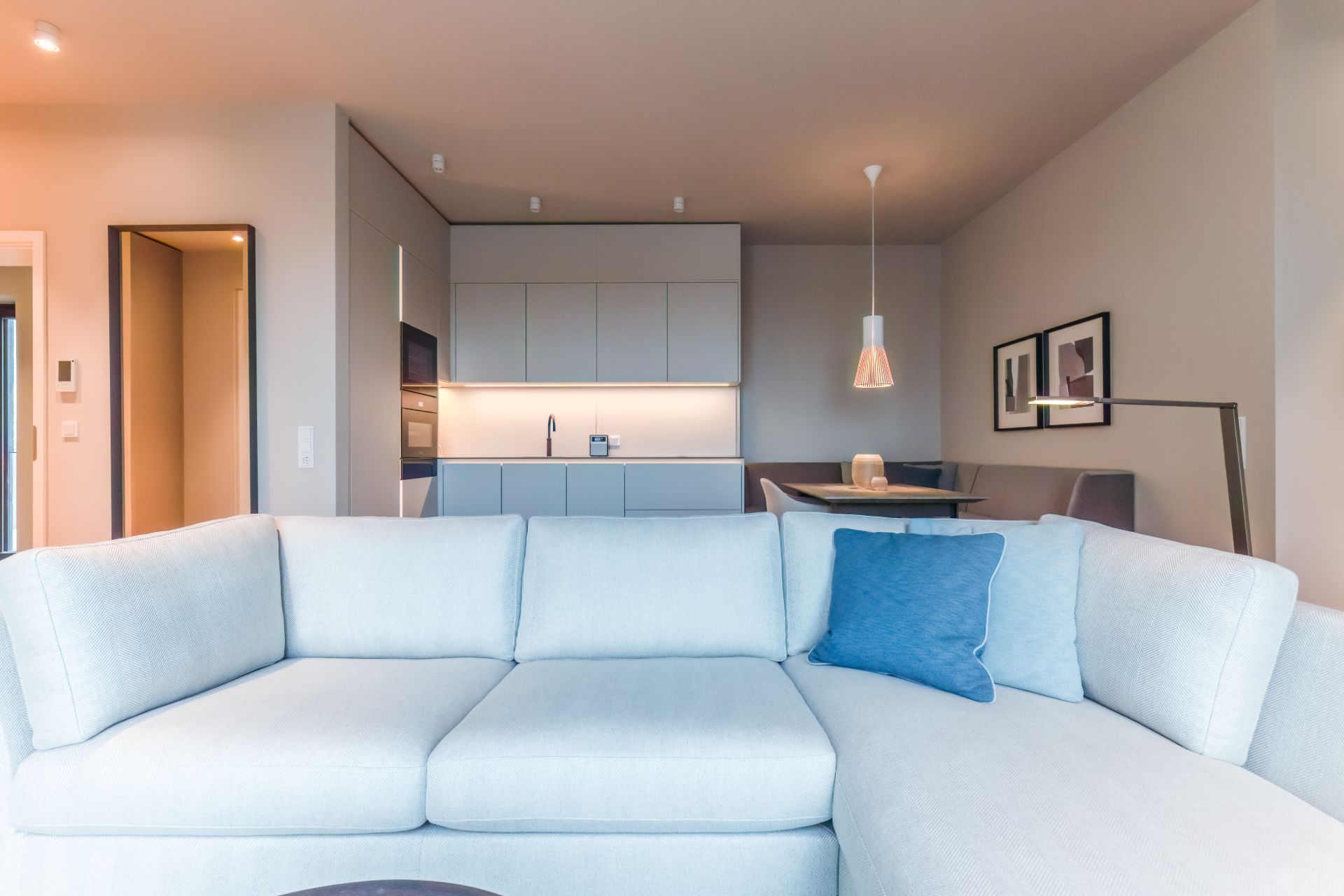 Helles Wohnzimmer mit Ecksofa, offener Küche und Esstisch mit Hängelampe.