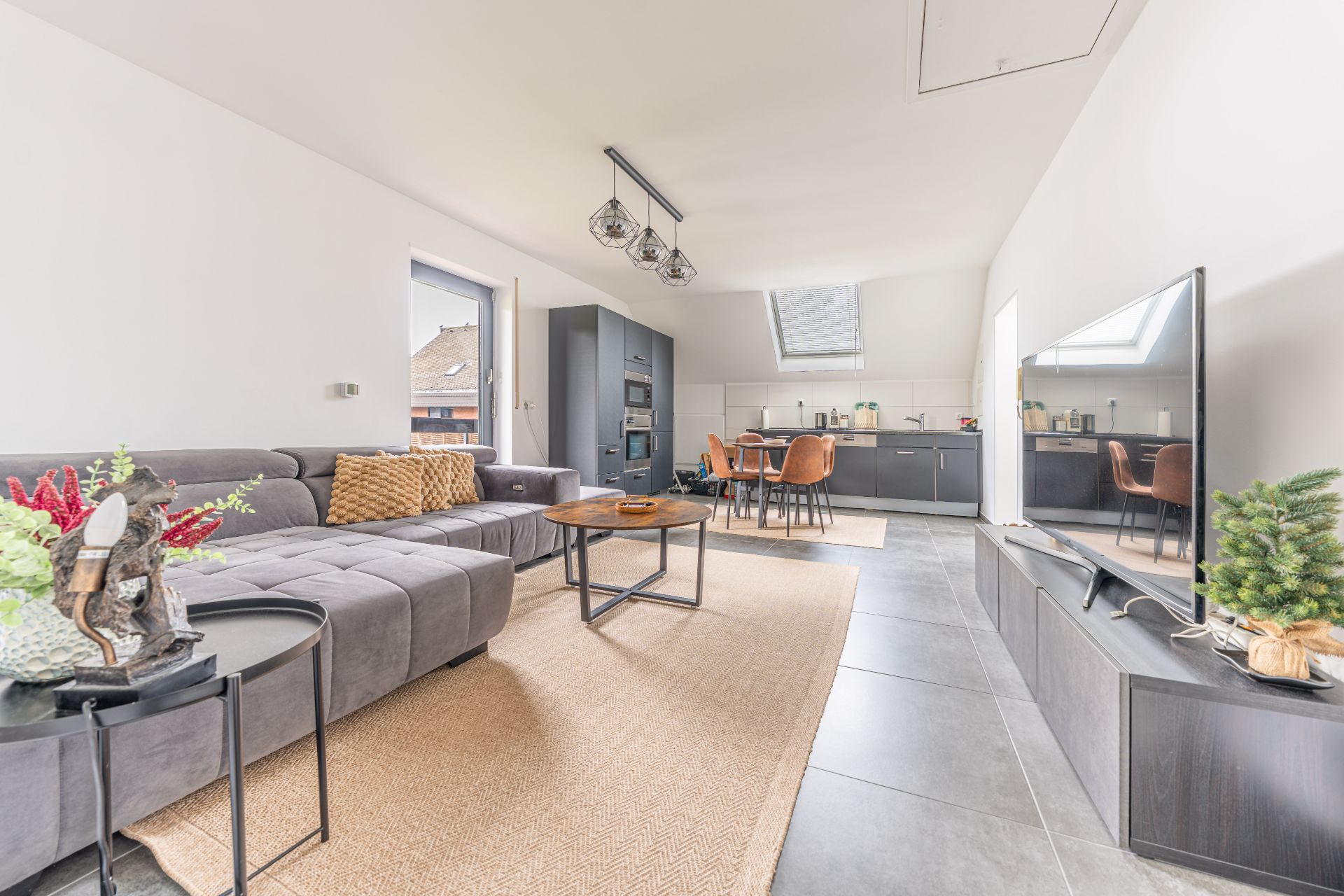 Helles Wohnzimmer mit grauem Sofa, Fernseher, Esstisch und offener Küche.