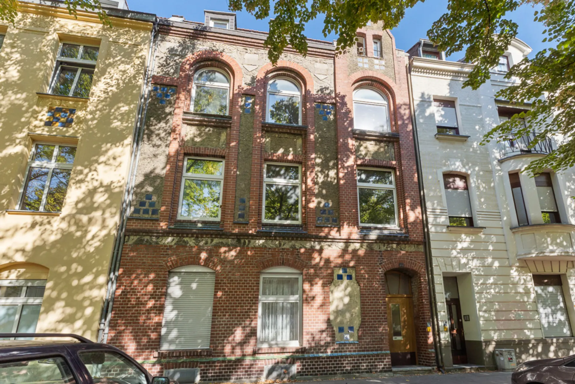 Dreistöckiges Wohnhaus mit roter Backsteinfassade und großen Fenstern, umgeben von Bäumen.