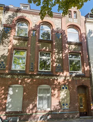 Dreistöckiges Wohnhaus mit roter Backsteinfassade und großen Fenstern, umgeben von Bäumen.