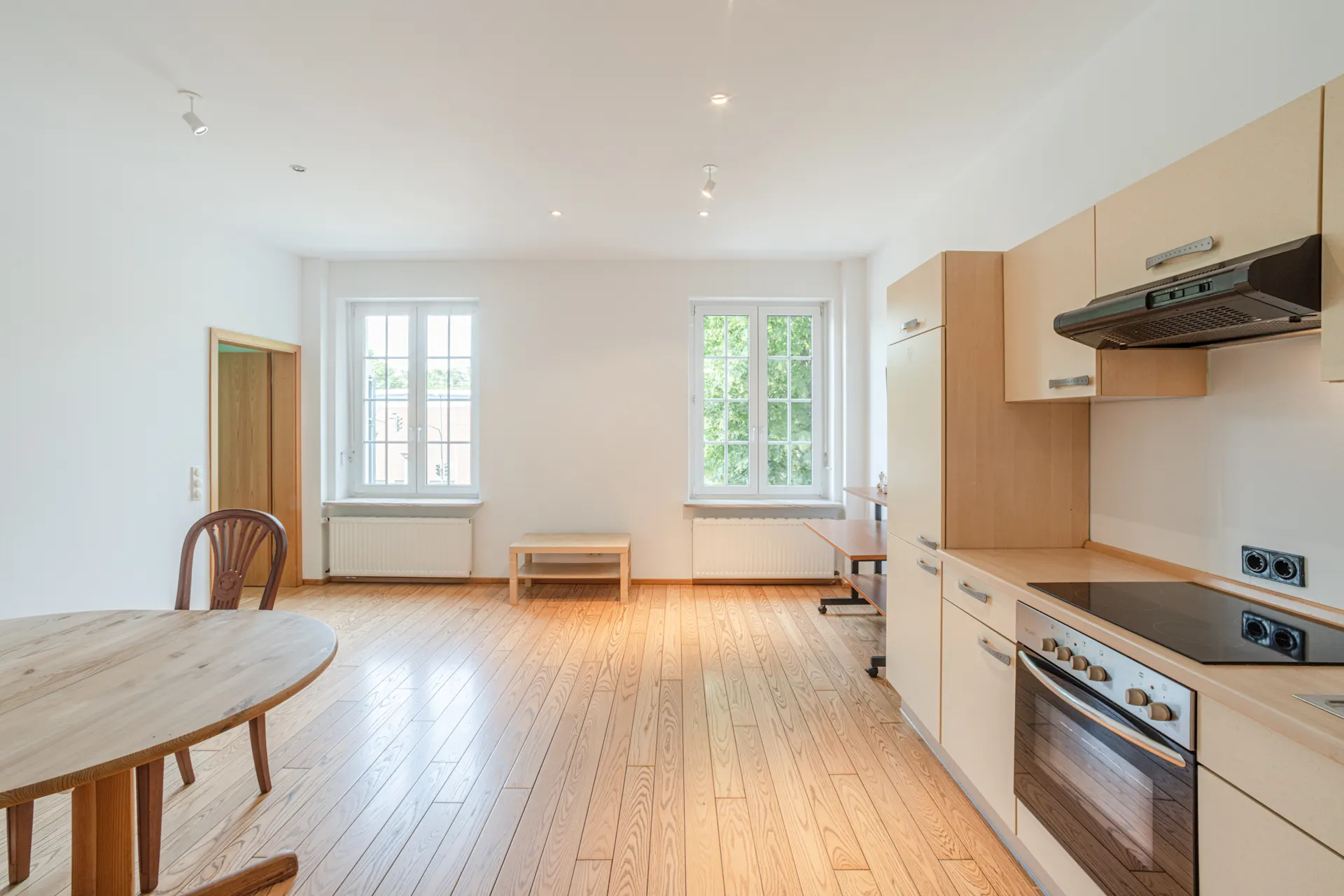 Helles Wohn- und Küchenzimmer mit Holzfußboden, Esstisch, Stühlen und zwei großen Fenstern.
