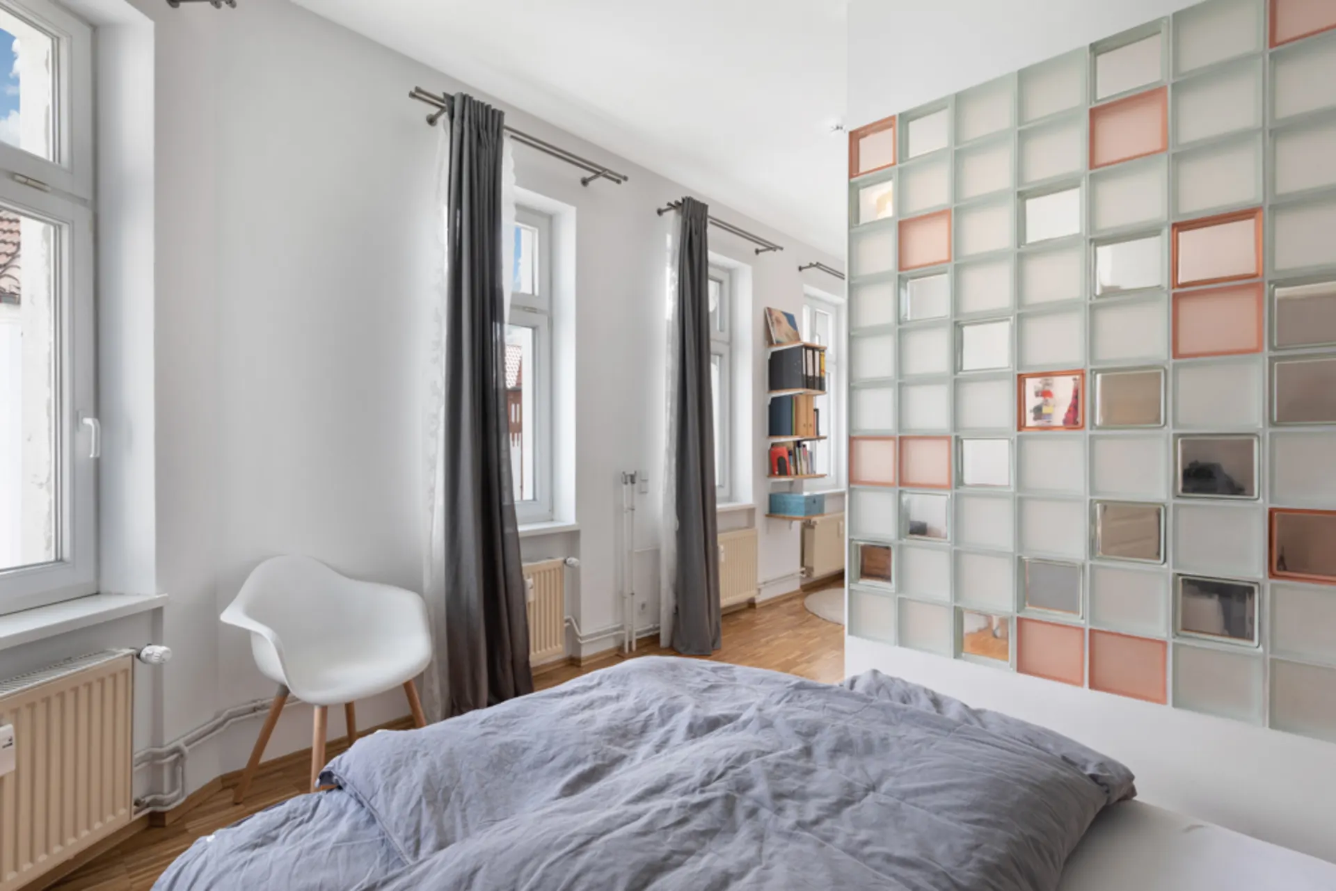 Helles Schlafzimmer mit Bett, Stuhl, großen Fenstern und einer Glasbausteinwand.