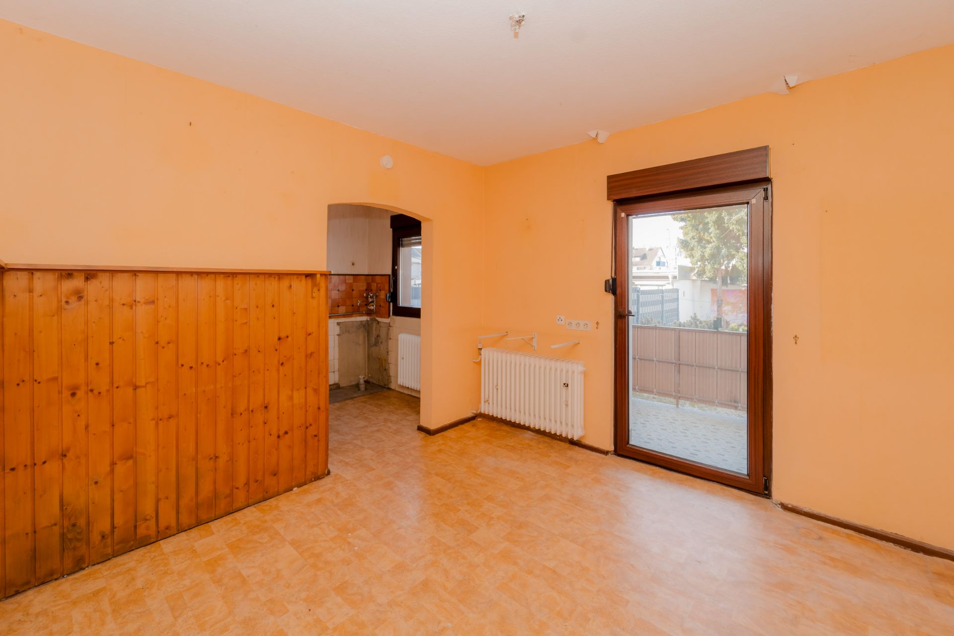 Leeres Zimmer mit orange gestrichenen Wänden, Holzwand, Tür zum Garten und Durchgang zur Küche.