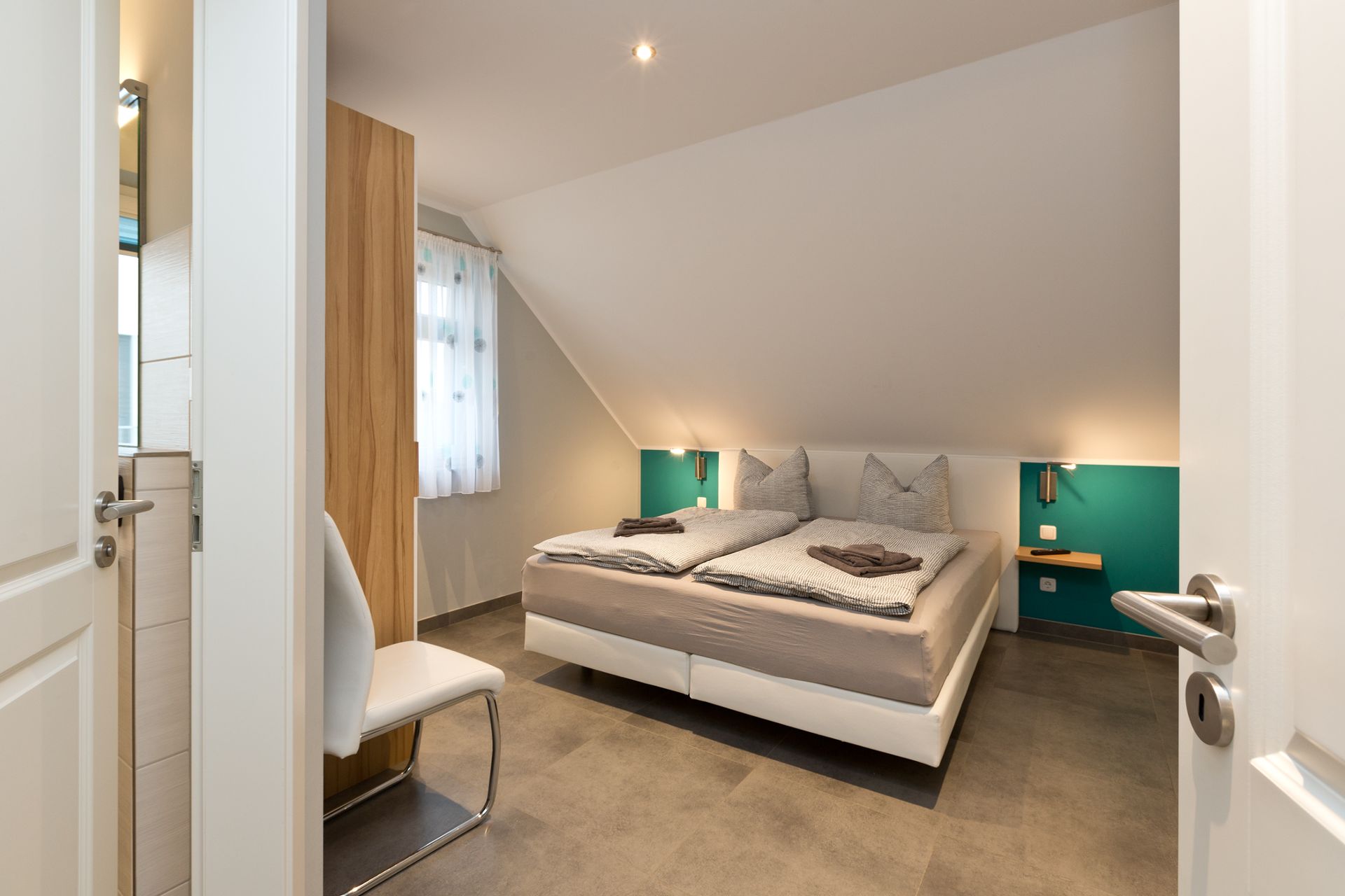 Modernes Schlafzimmer mit Doppelbett unter einer Dachschräge, seitlich ein Fenster und ein Stuhl.