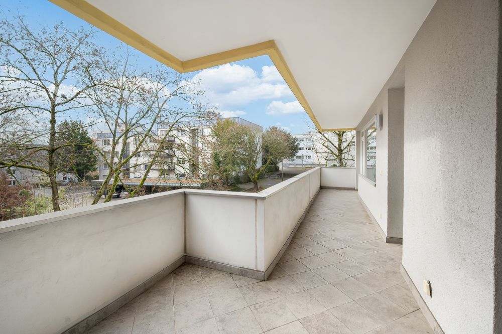 Großer, überdachter Balkon mit Fliesenboden und Blick auf Bäume und Wohngebäude.
