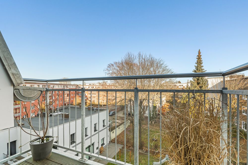 Balkon mit Metallgeländer, zwei Pflanzen, Blick auf Wohngebäude und Bäume bei klarem Himmel.