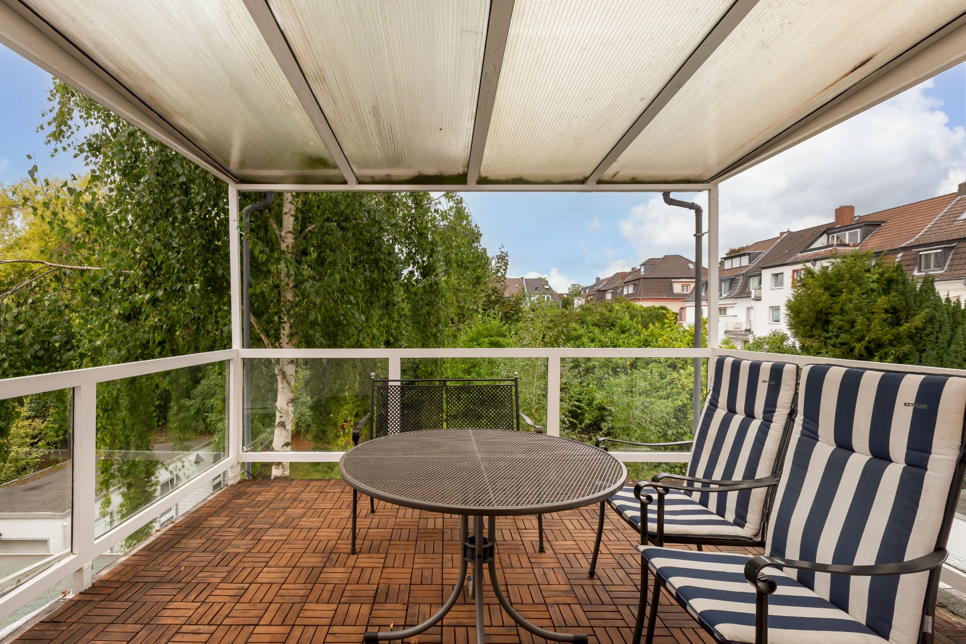 Überdachte Terrasse mit Holzboden, rundem Tisch und gestreiften Stühlen, Blick auf Bäume und Häuser.