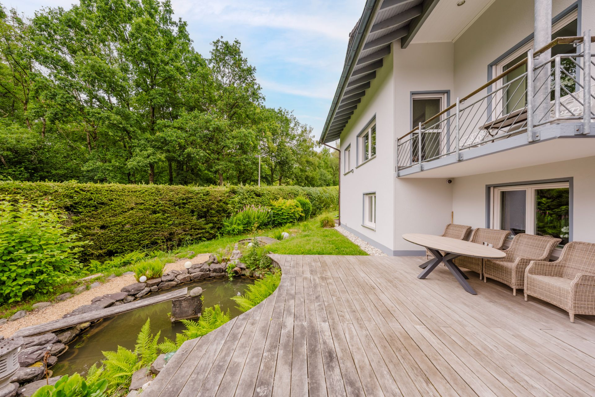 Große Holzterrasse mit Gartenmöbeln neben einem kleinen Teich und viel Grün.