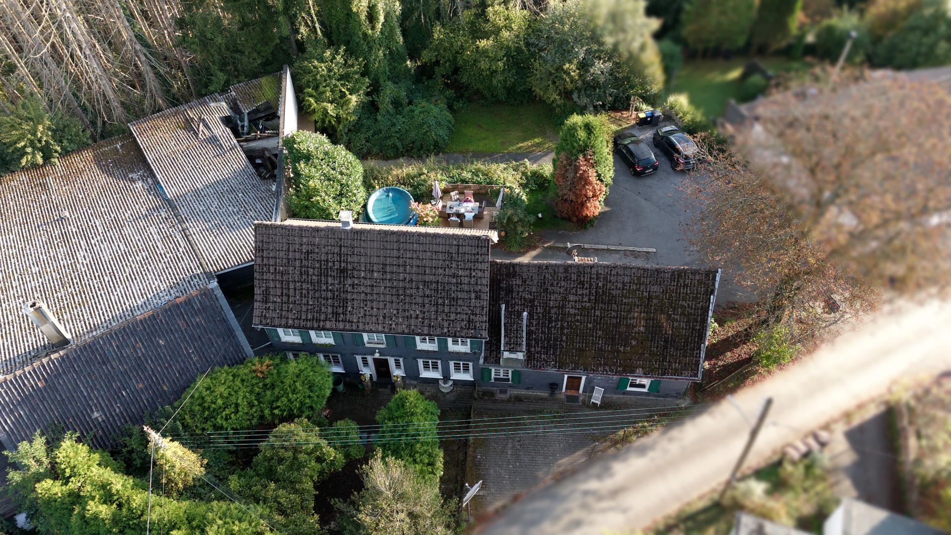 Luftaufnahme eines Wohnhauses mit Garten, kleinem Pool und geparkten Autos in einer grünen Umgebung.