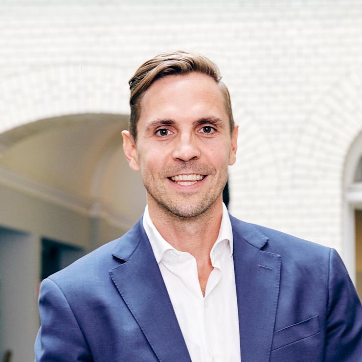 Ein Mann im blauen Anzug steht in einem Innenhof mit weißen Backsteinwänden.