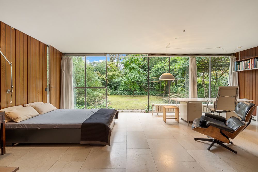 Modernes Schlafzimmer mit großen Fenstern, Blick auf Garten, Schreibtisch und Sessel.