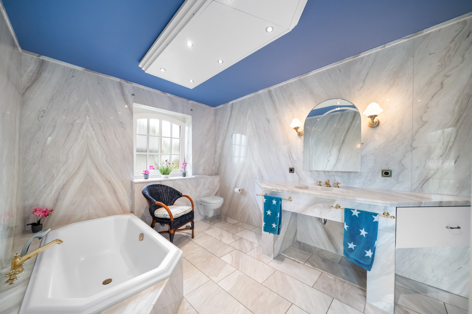 Helles Badezimmer mit Badewanne, Doppelwaschbecken, Fenster, Stuhl und blauem Deckenanstrich.