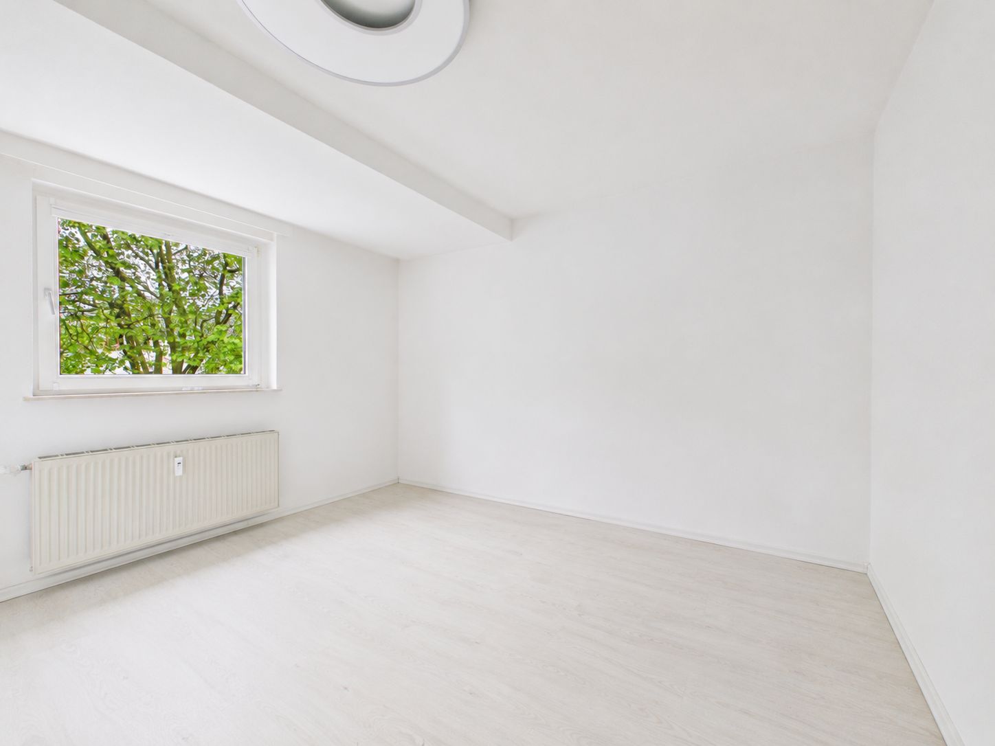 Leerer, heller Raum mit weißem Boden, Fenster und Heizkörper an der Wand.