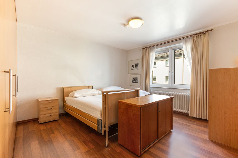 Schlafzimmer mit Holzfußboden, zwei Einzelbetten, Schrank, Nachttisch und großem Fenster mit Vorhängen.