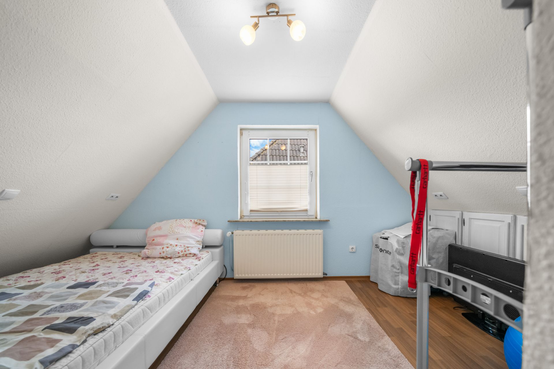 Kleines Dachzimmer mit Einzelbett, Fenster, Teppich und Tageslichtlampe.