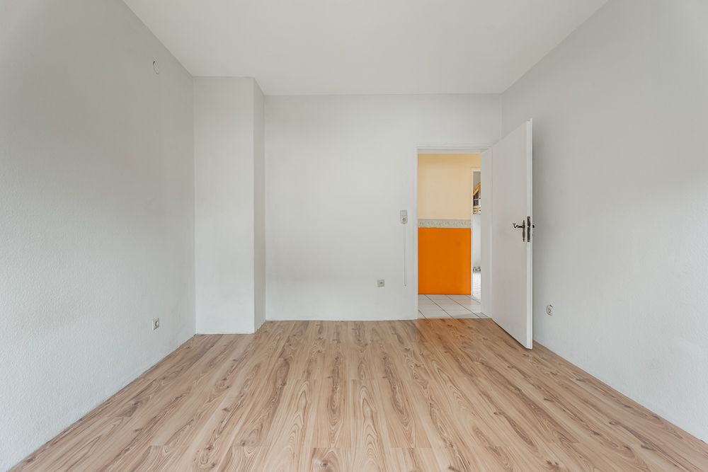 Leerer Raum mit hellem Laminatboden und weißer Tür zur angrenzenden Küche mit orangefarbener Verkleidung.