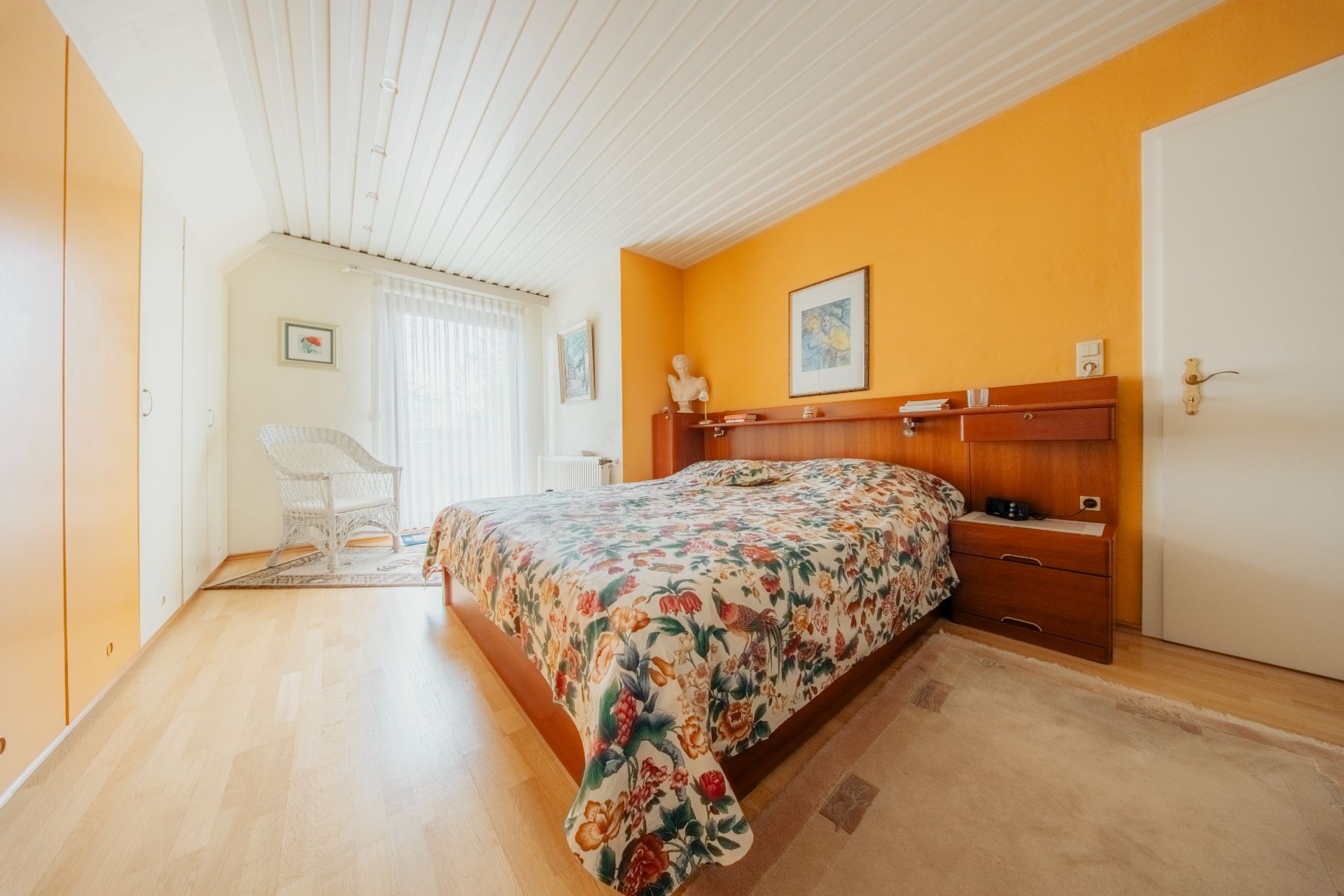 Helles Schlafzimmer mit Doppelbett, Blumenbettdecke, gelber Wand und Korbstuhl am Fenster.