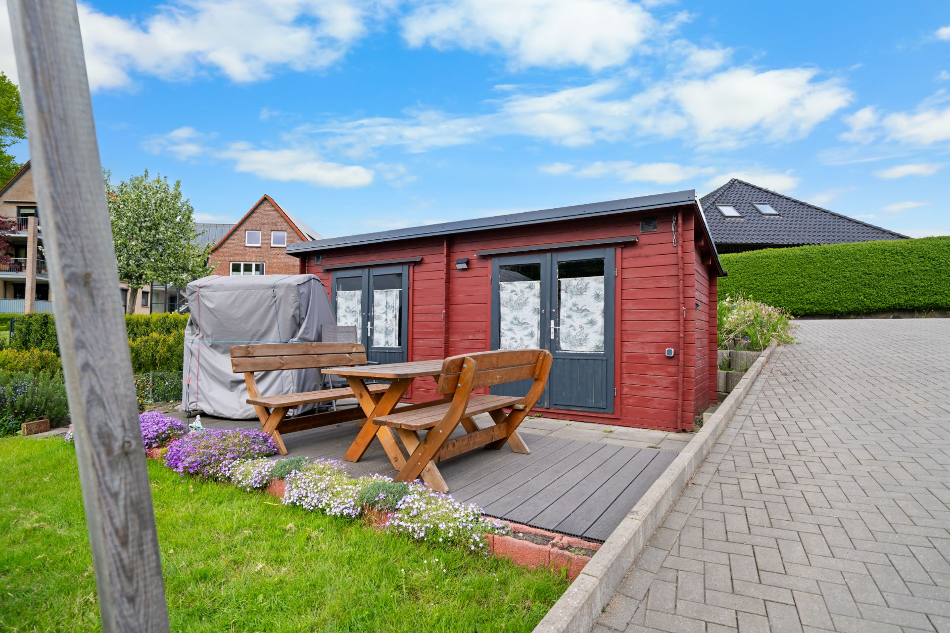 Kleines rotes Gartenhaus mit Holzbänken und Tisch auf einer Terrasse im Grünen.