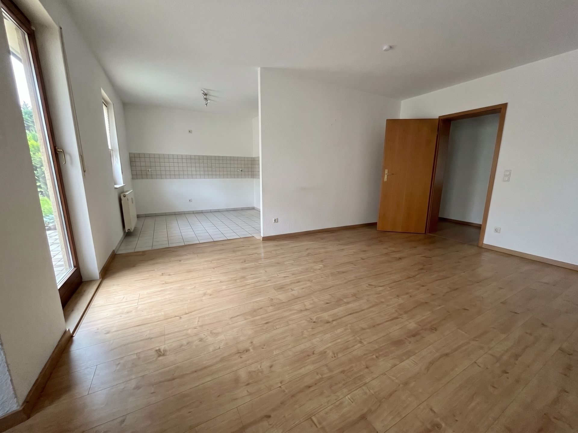 Leerer Wohnraum mit Holzboden, offener Küche und einer Tür in einen weiteren Raum.