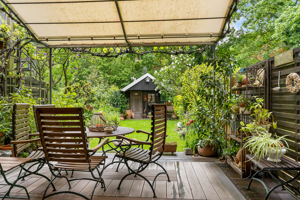 Überdachte Terrasse mit Holztisch und Stühlen, Pflanzen und Blick auf einen Garten mit Gartenhaus.