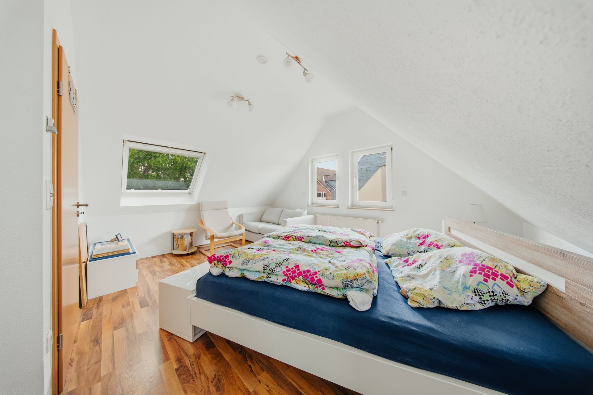 Helles Schlafzimmer mit Doppelbett, bunter Bettwäsche, Sessel und zwei Fenstern unter Dachschräge.