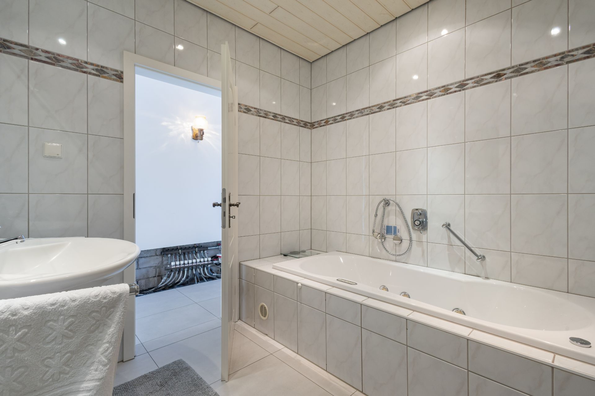 Helles Badezimmer mit gefliester Badewanne, Waschbecken und offener Tür zu einem weiteren Raum.