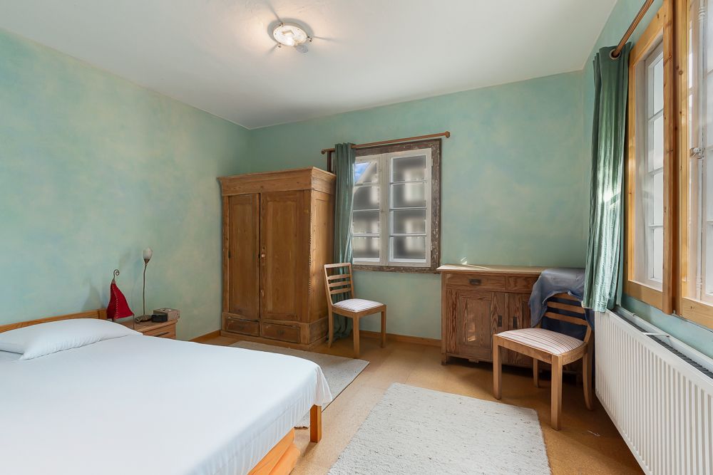 Schlafzimmer mit Bett, Holzschrank, zwei Stühlen, Schreibtisch und großen Fenstern.