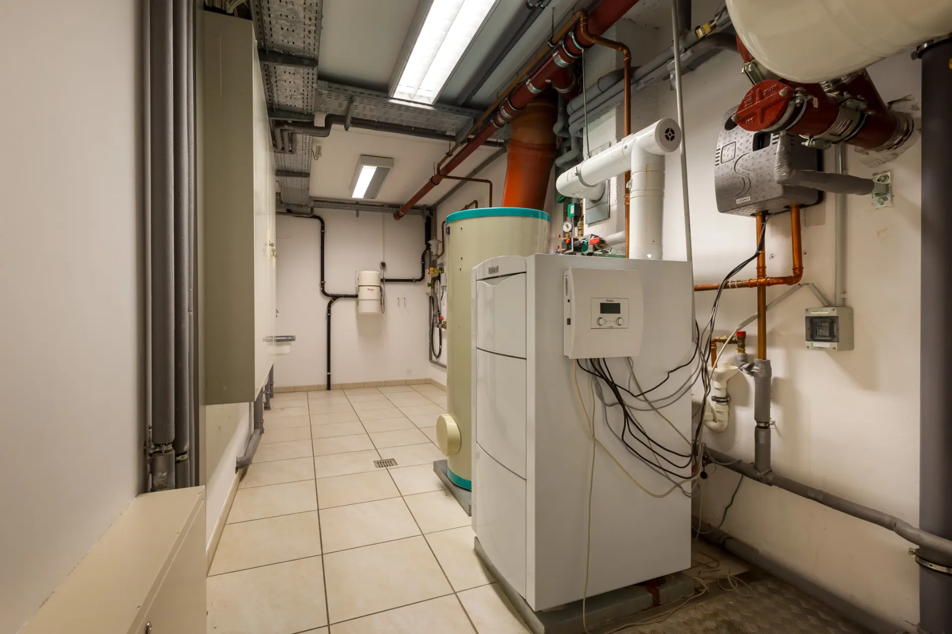 Heizungsraum mit Boiler, Rohren und technischen Anlagen, gefliestem Boden und weißen Wänden.
