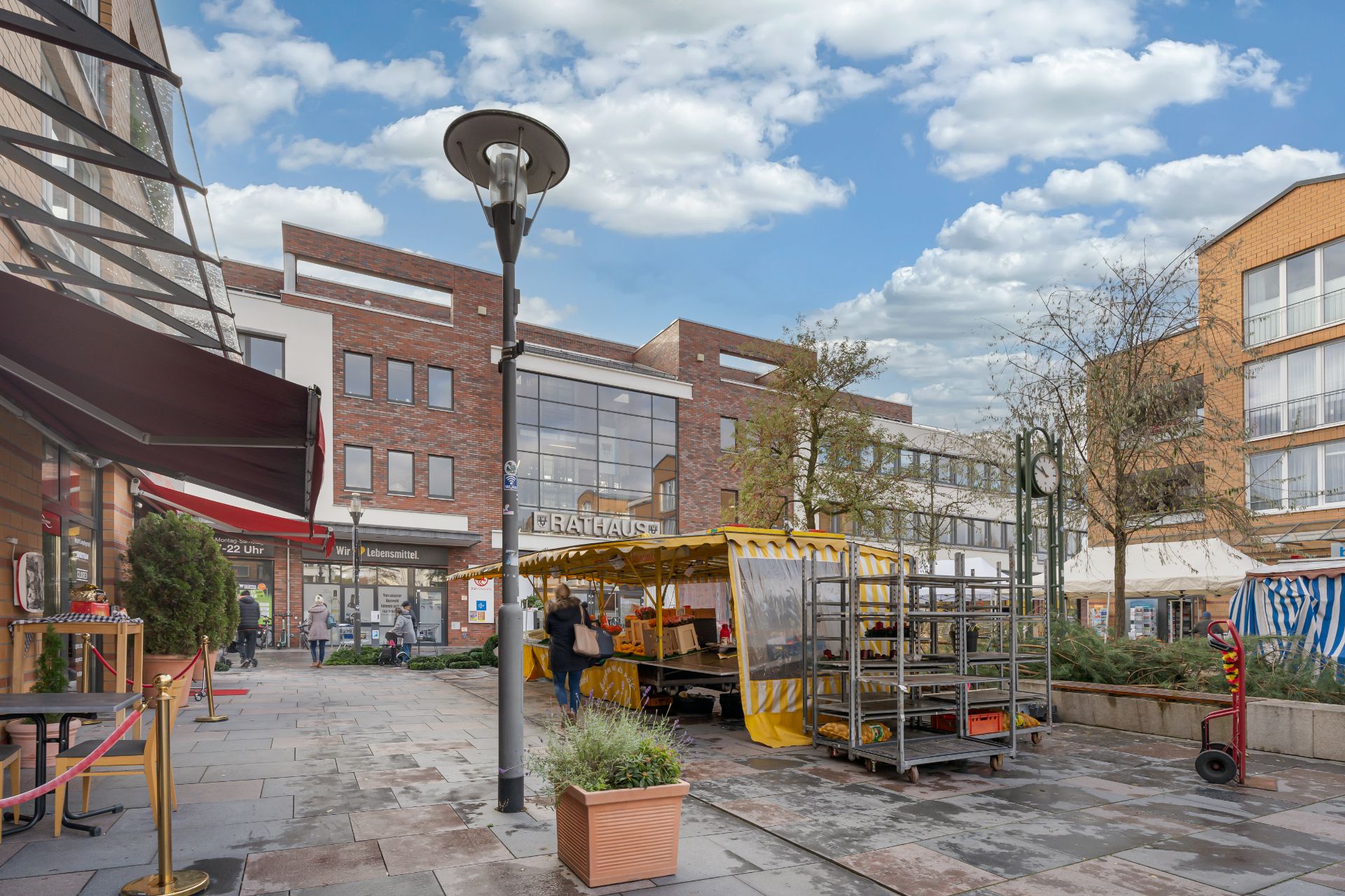 Stadtplatz mit Marktständen, Menschen, Laternen und modernen Gebäuden im Hintergrund.