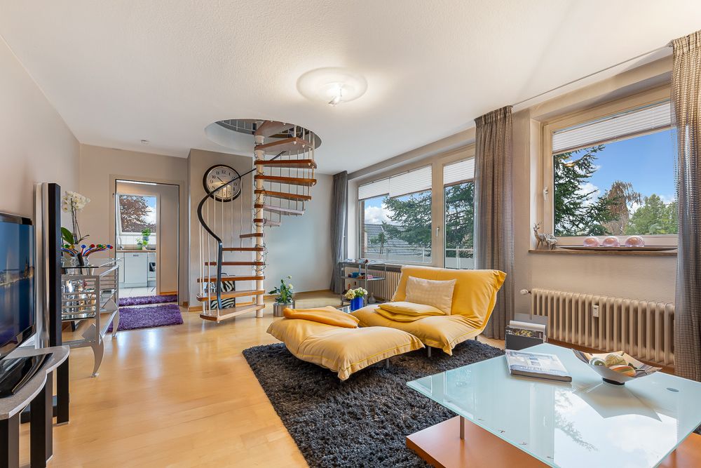 Modernes Wohnzimmer mit gelbem Sofa, Glastisch und Wendeltreppe, große Fenster mit Blick ins Grüne.