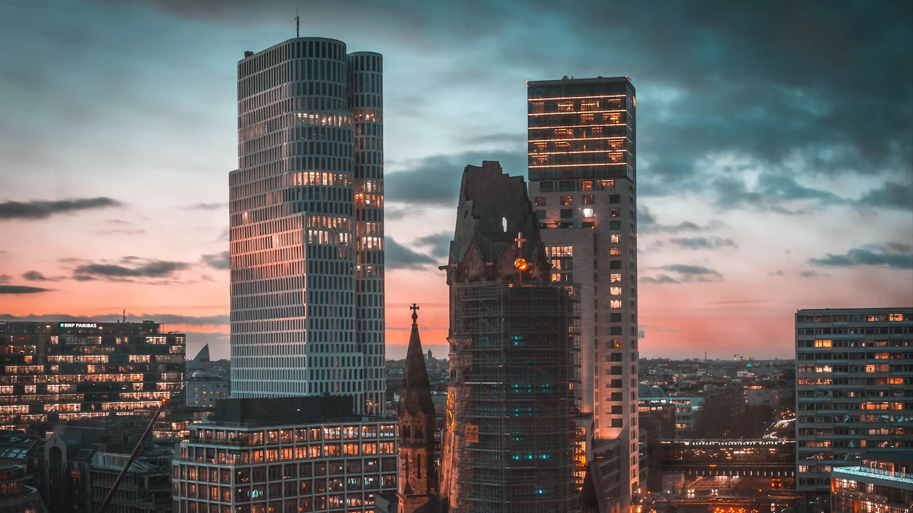 Stadtansicht mit modernen Hochhäusern und einer historischen Kirche im Vordergrund bei Sonnenuntergang.