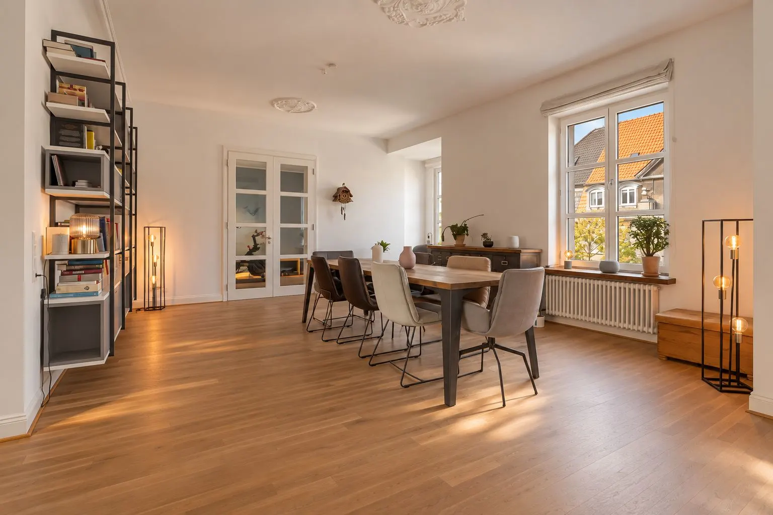 Helles Esszimmer mit großem Holztisch, sechs Stühlen und Regal, Holzboden und Fenster.