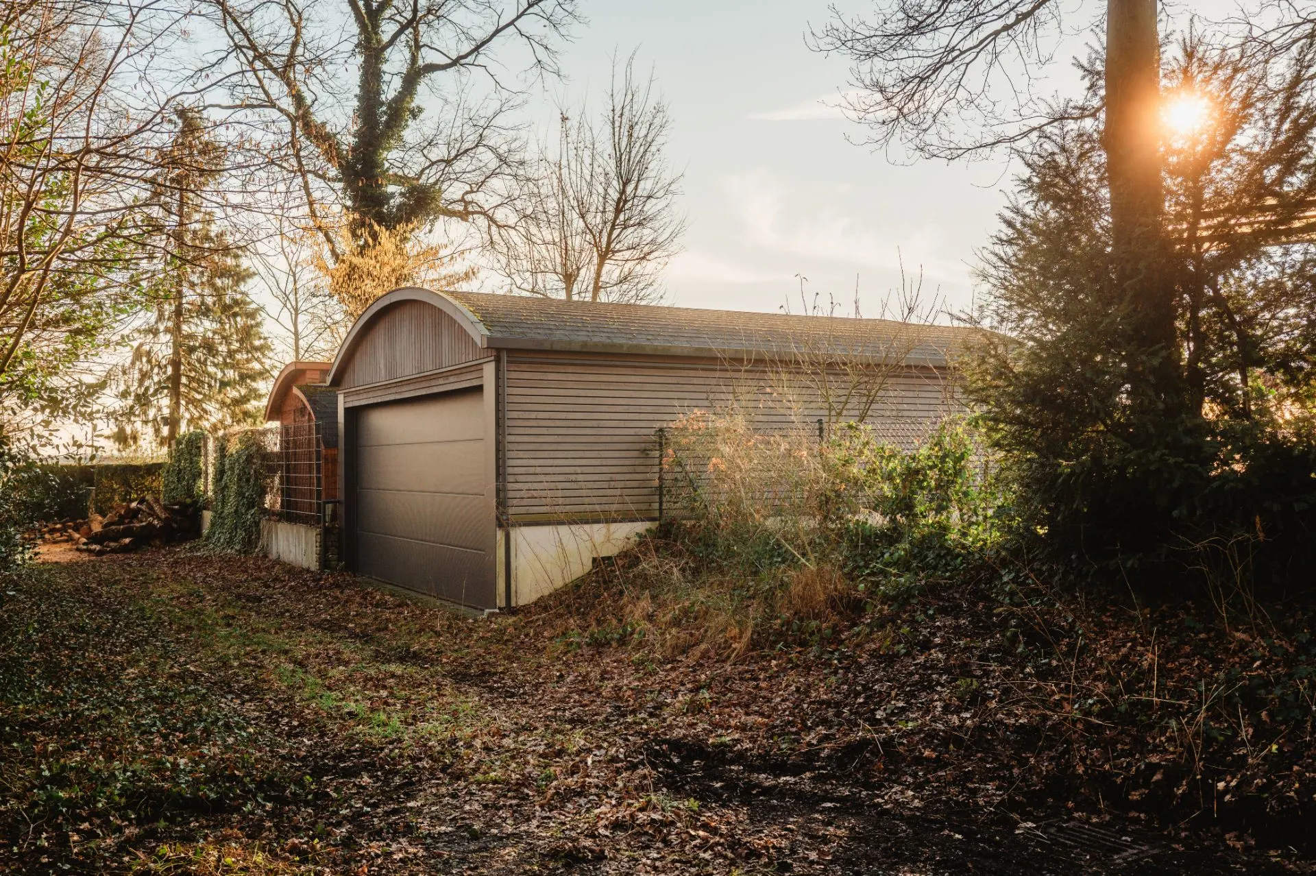 Eine Holzgarage mit rundem Dach steht an einem bewachsenen Grundstück, umgeben von Bäumen.