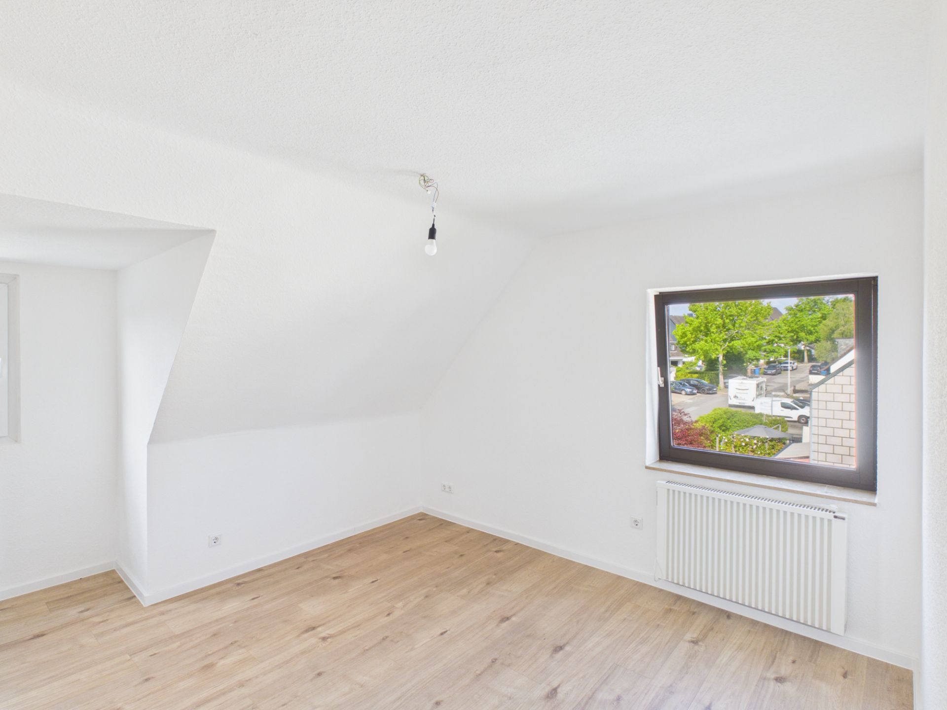 Helles, leeres Zimmer mit Holzfußboden, weißer Wand und großem Fenster mit Blick nach draußen.