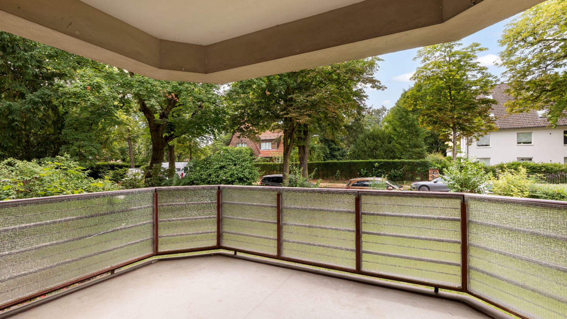 Balkon mit Glasgeländer, Blick auf Garten, Bäume und geparkte Autos vor Wohnhäusern.