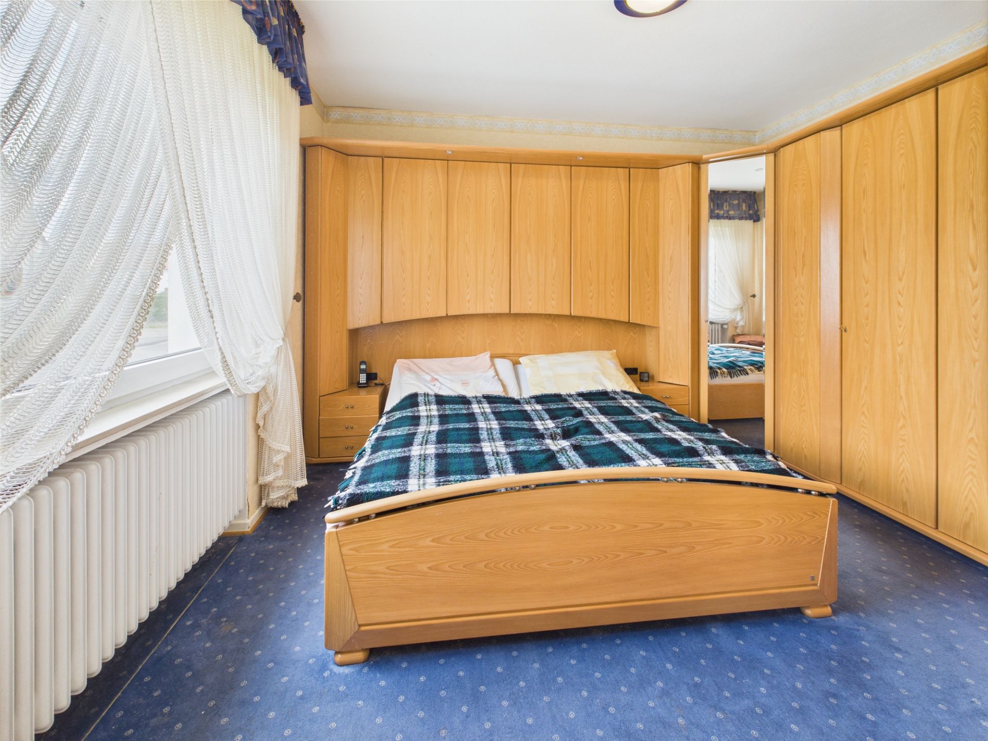 Schlafzimmer mit Holzbett, großer Eckschrankwand und Fenster mit weißen Vorhängen.