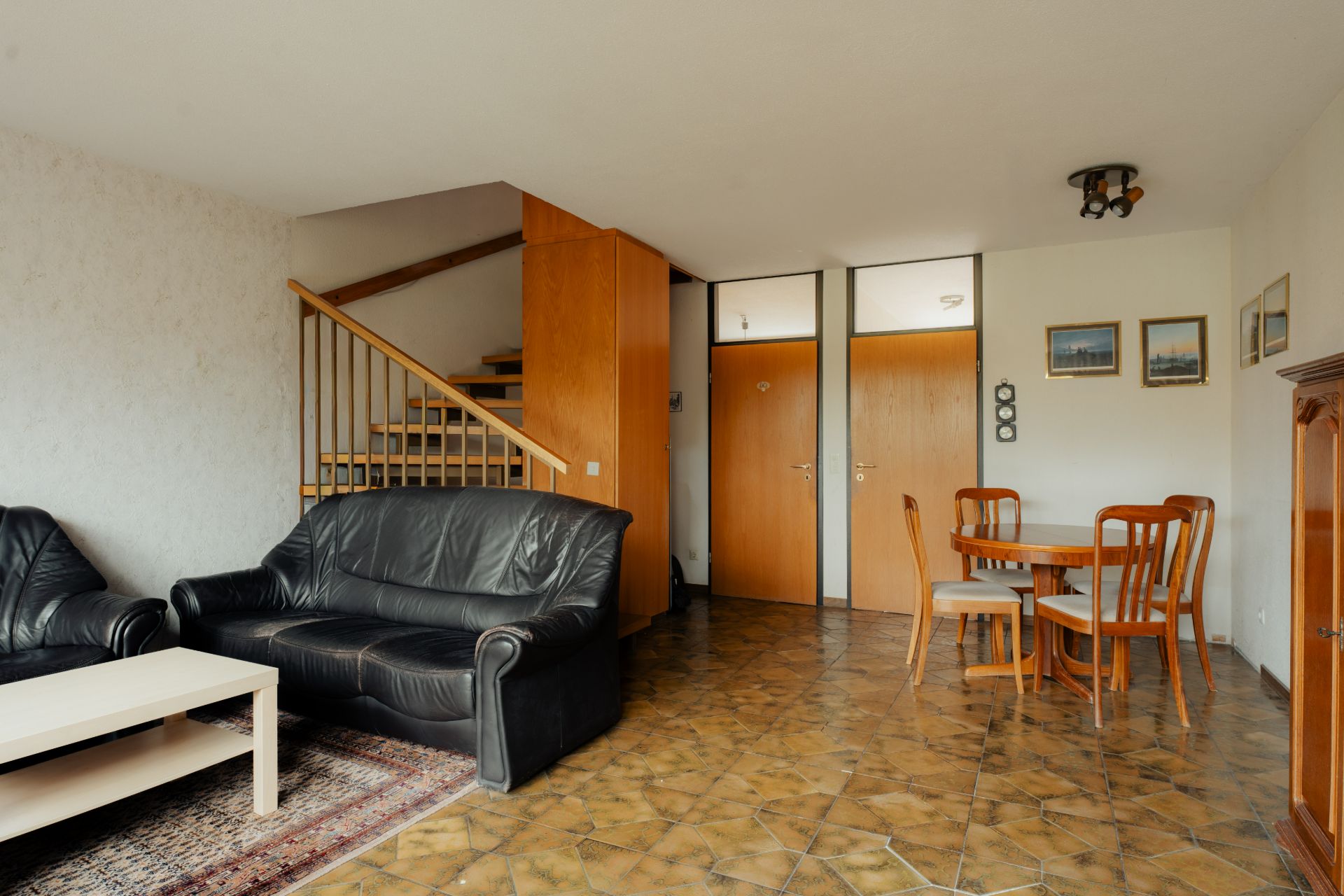 Wohnzimmer mit schwarzem Ledersofa, Esstisch mit Stühlen und Treppe im Hintergrund.