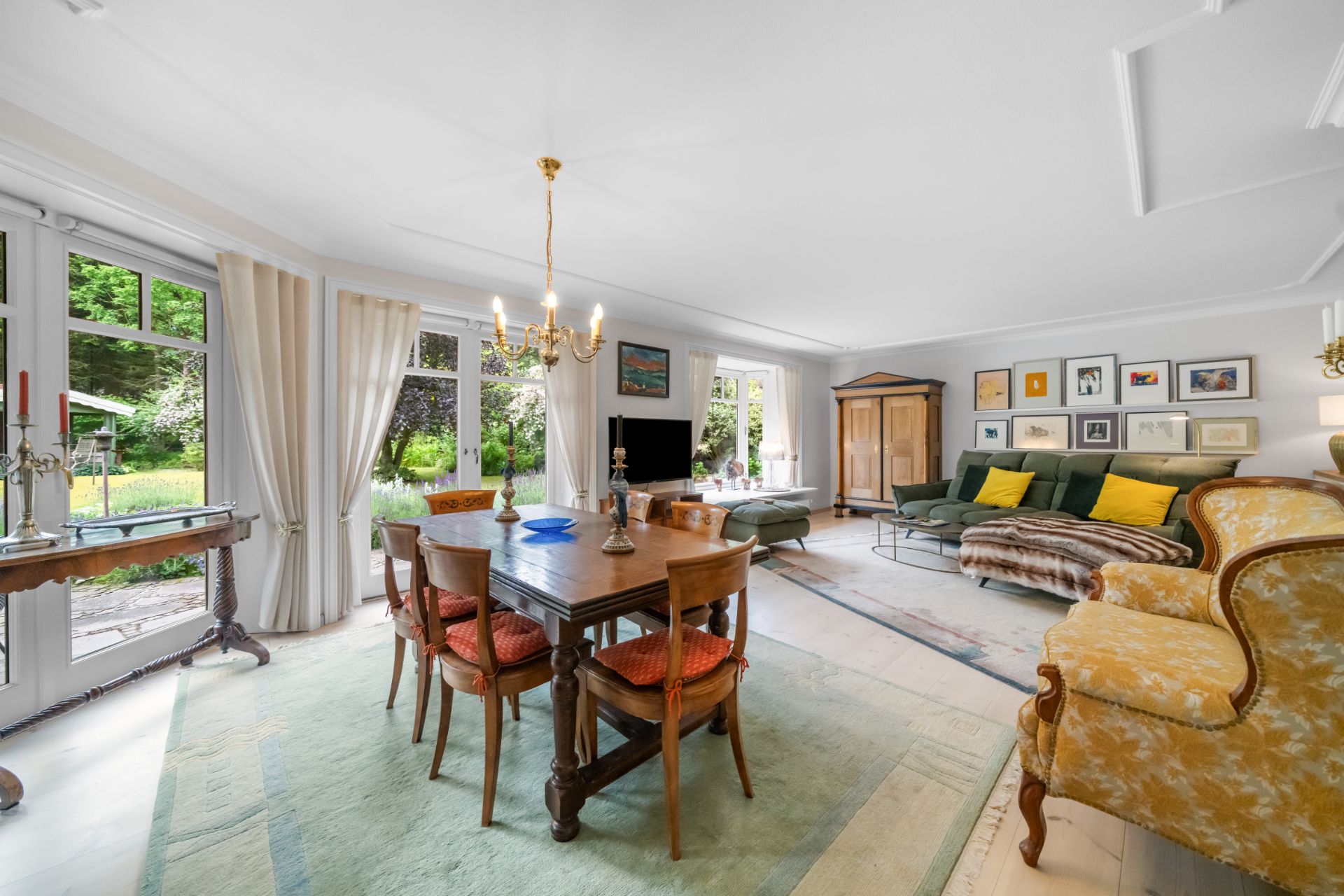 Helles Wohnzimmer mit Esstisch, Sesseln, großer Couch, Bildern an der Wand und Blick in den Garten.