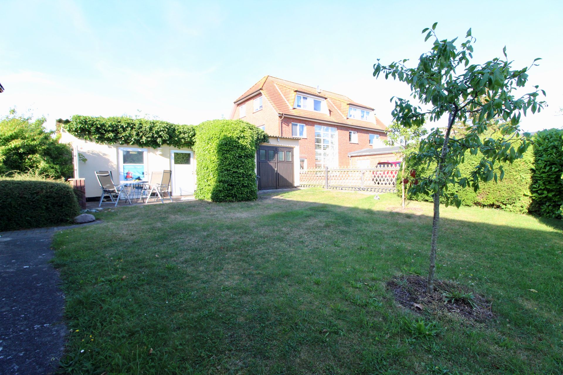 Grüner Garten mit kleinem Baum, Rasenfläche und Terrasse mit Gartenmöbeln vor einem Wohnhaus.