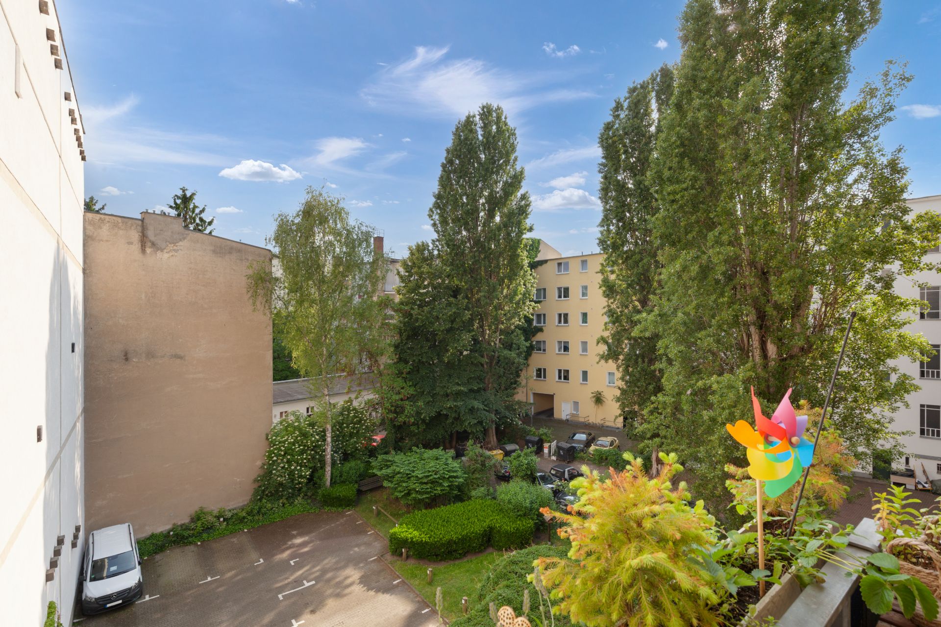 Blick auf einen begrünten Innenhof mit Bäumen, Sträuchern und einem bunten Windrad auf dem Balkon.