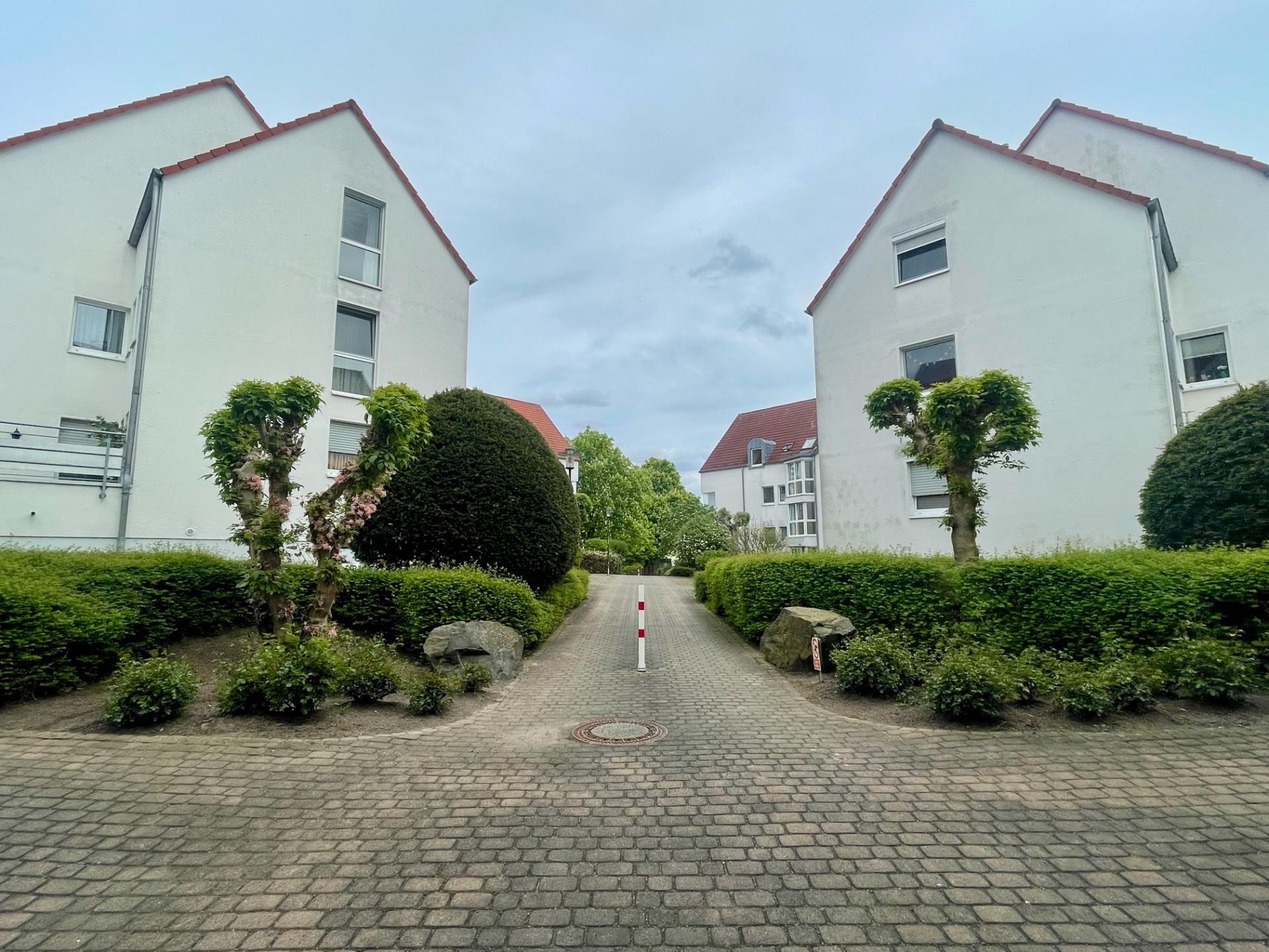 Gepflasterter Weg zwischen zwei weißen Wohngebäuden mit Büschen und Bäumen an den Seiten.