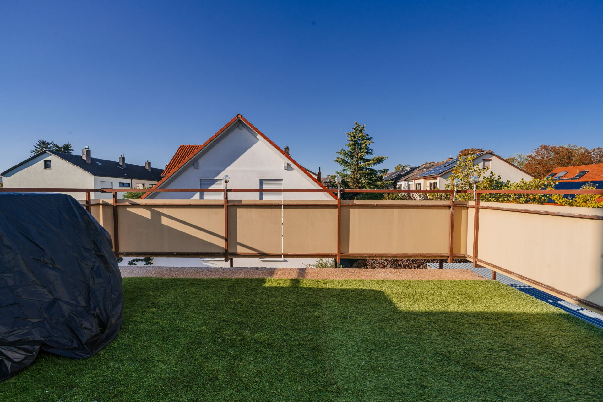Blick von einer Terrasse mit grünem Rasen und Zaun auf Nachbarhäuser unter blauem Himmel.