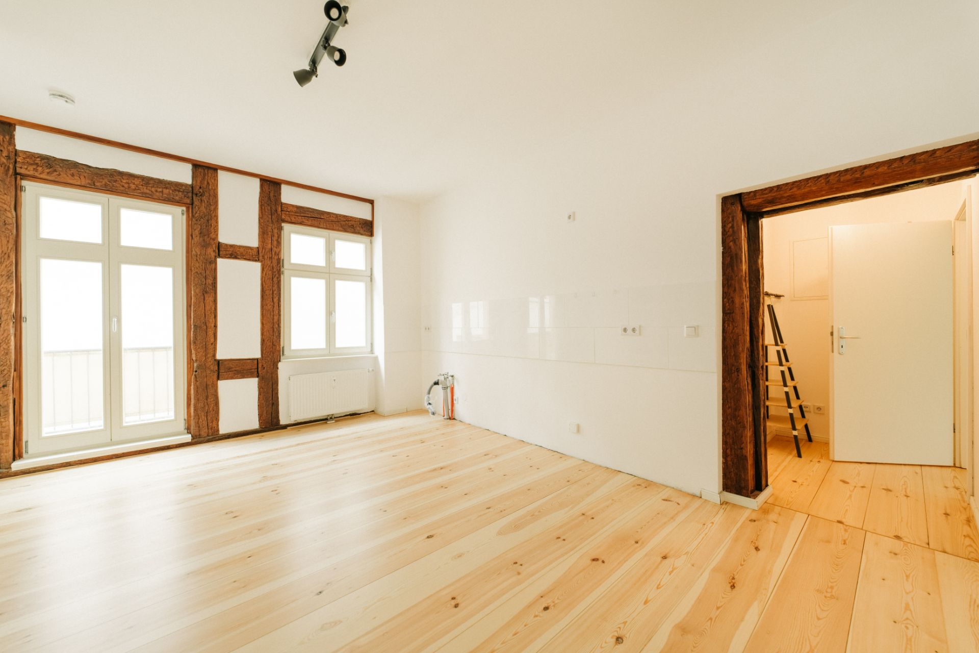Heller Raum mit Holzboden, großen Fenstern, sichtbaren Holzbalken und einer offenen Tür zu einem weiteren Raum.