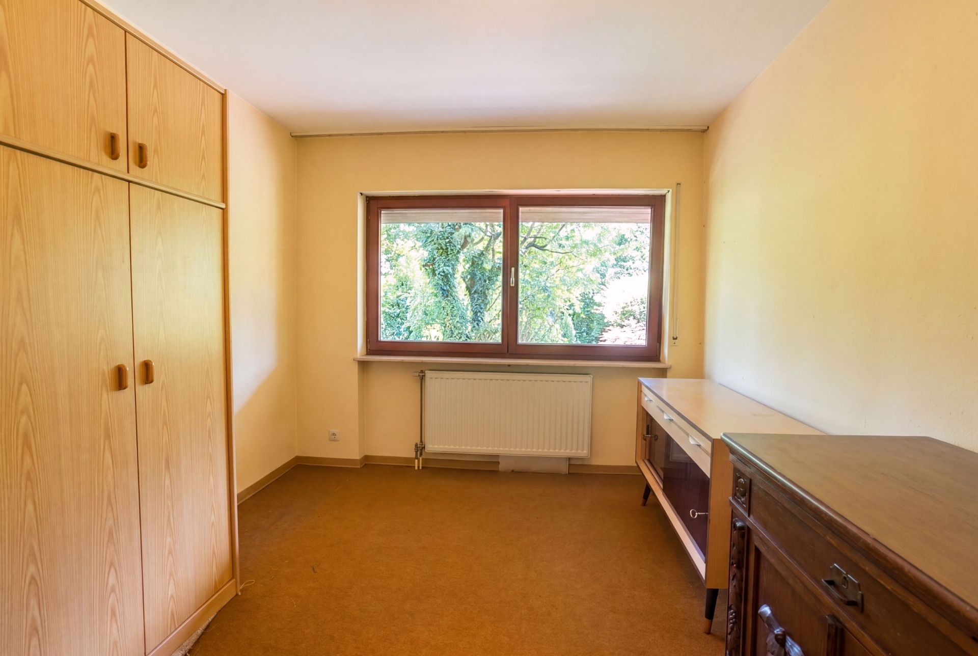 Helles Zimmer mit großem Fenster, Kleiderschrank, Schreibtisch und Kommode.