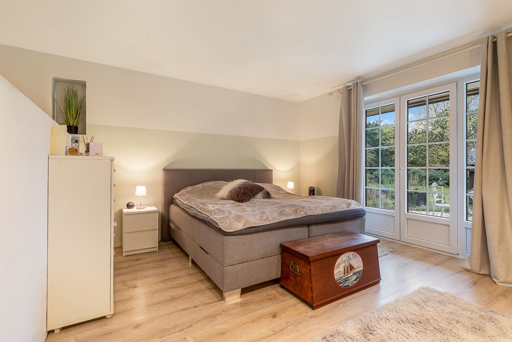 Helles Schlafzimmer mit Bett, Kommode, Truhe und großen Fenstern mit Blick ins Grüne.
