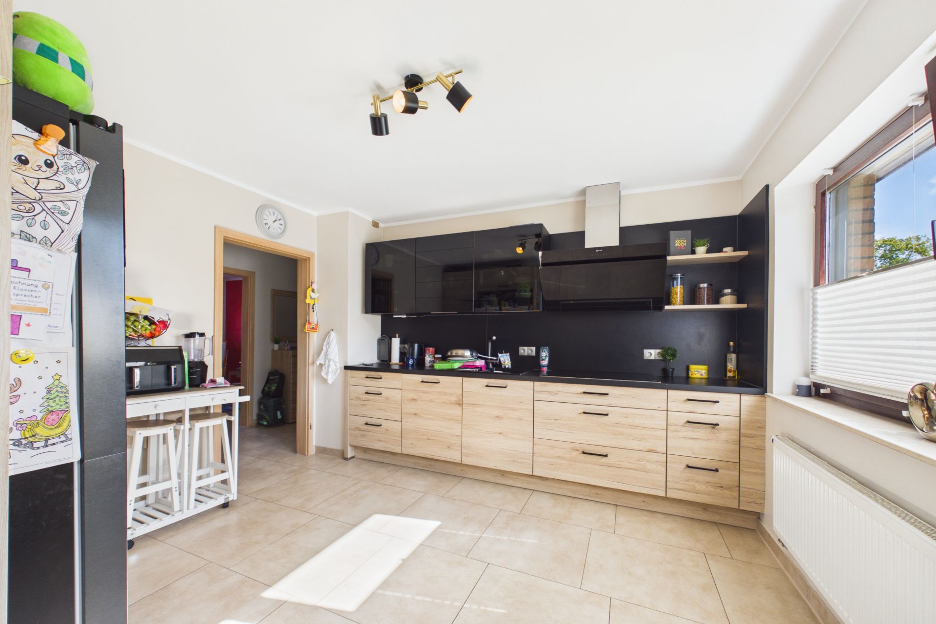 Moderne Küche mit Holzfronten, schwarzen Oberschränken und großen Fenstern, helle Fliesen am Boden.
