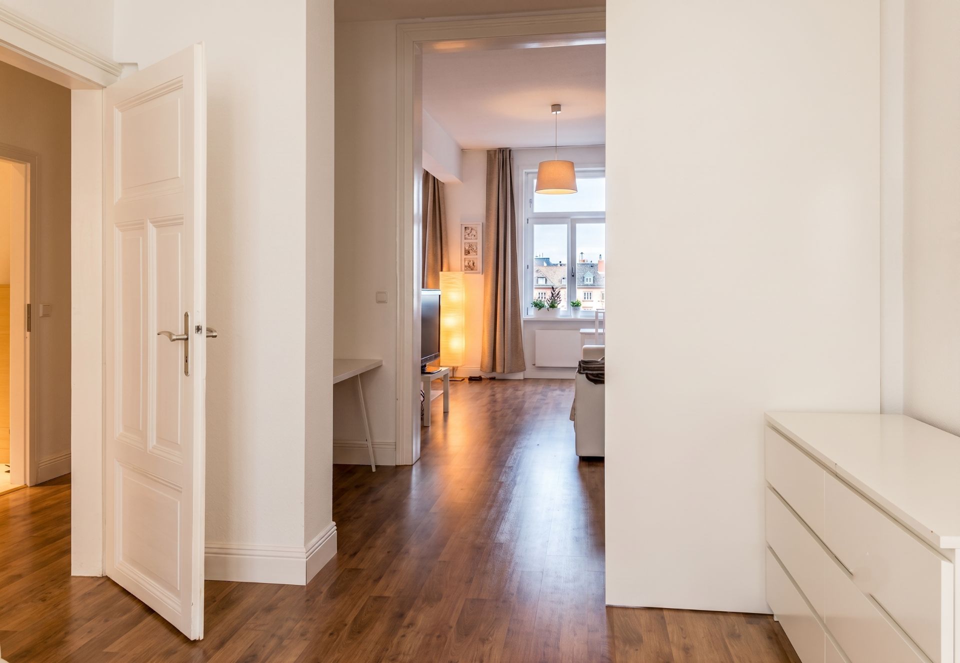 Heller Flur mit Holzboden, Blick ins Wohnzimmer mit großen Fenstern und Lampe.