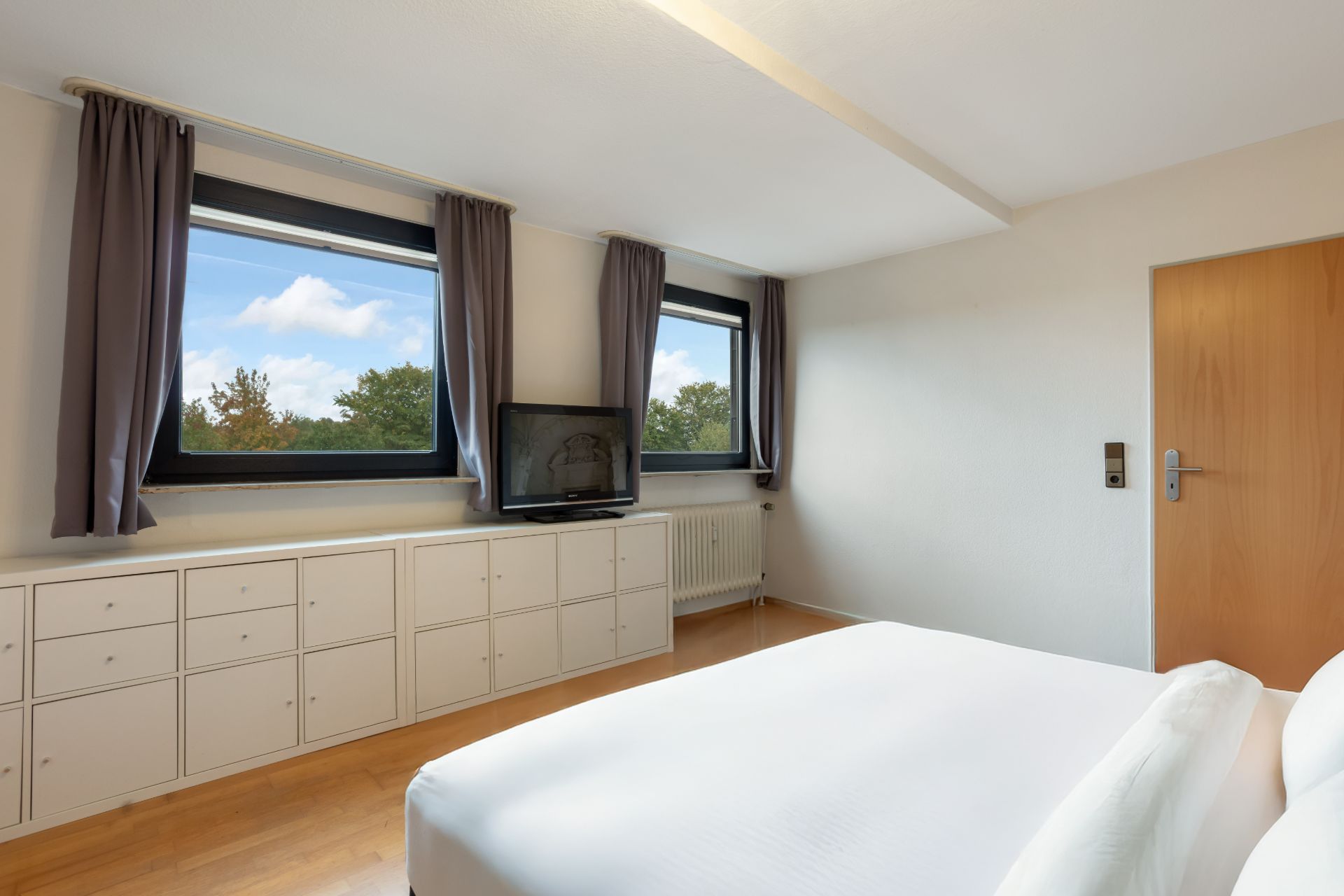 Helles Schlafzimmer mit zwei Fenstern, weißem Bett, TV und Kommode mit vielen Schubladen.